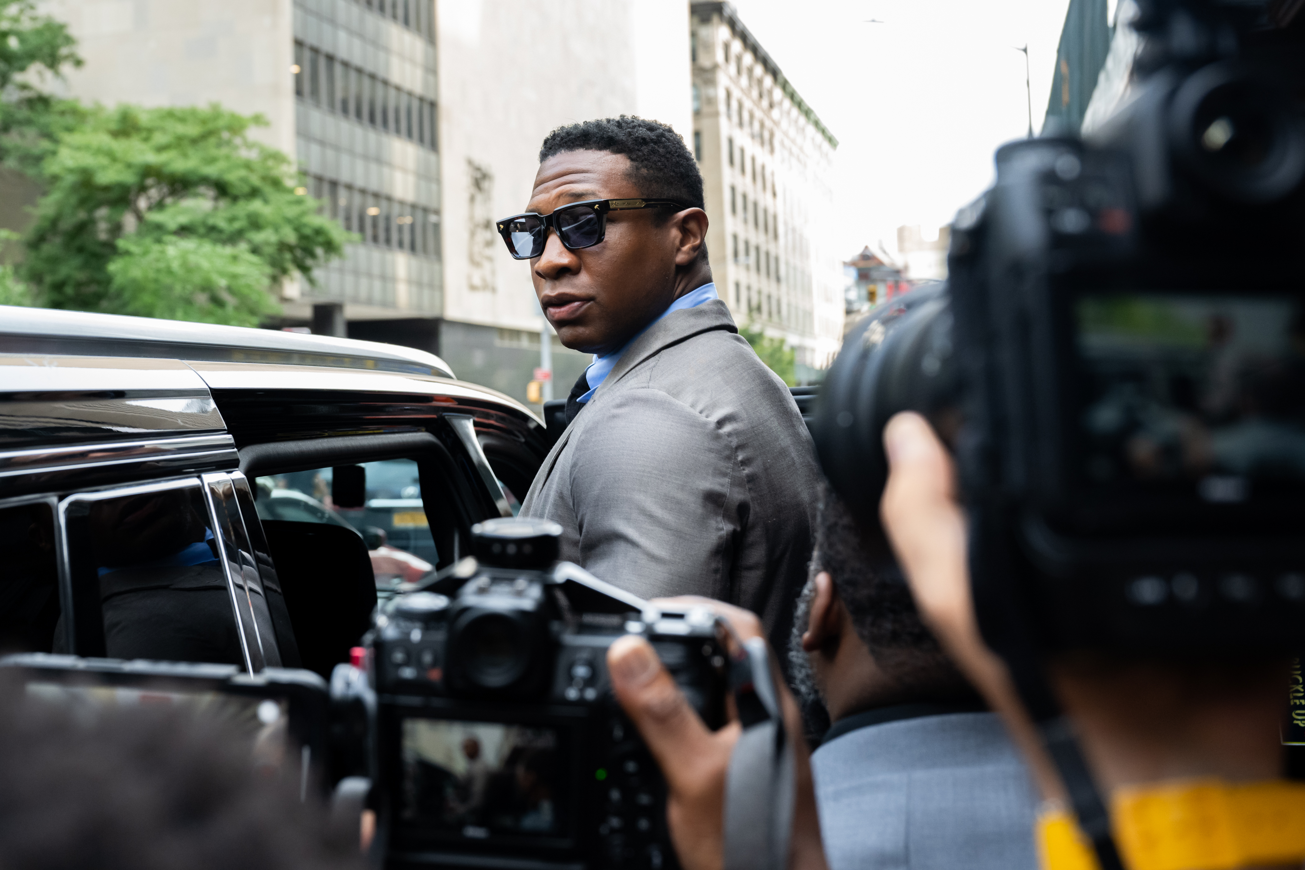 Jonathan Majors, in sunglasses and a grey suit, looks back over his shoulder at media cameras while getting into a car outside of Manhattan Criminal court after his pre-trial hearing on August 03, 2023 in New York City. 