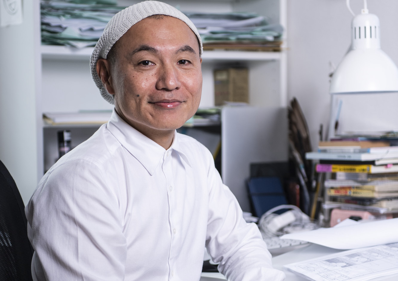Masaaki Yuasa in a studio smiling at the camera in a beanie and white shirt with storyboard pages on the desk beside him