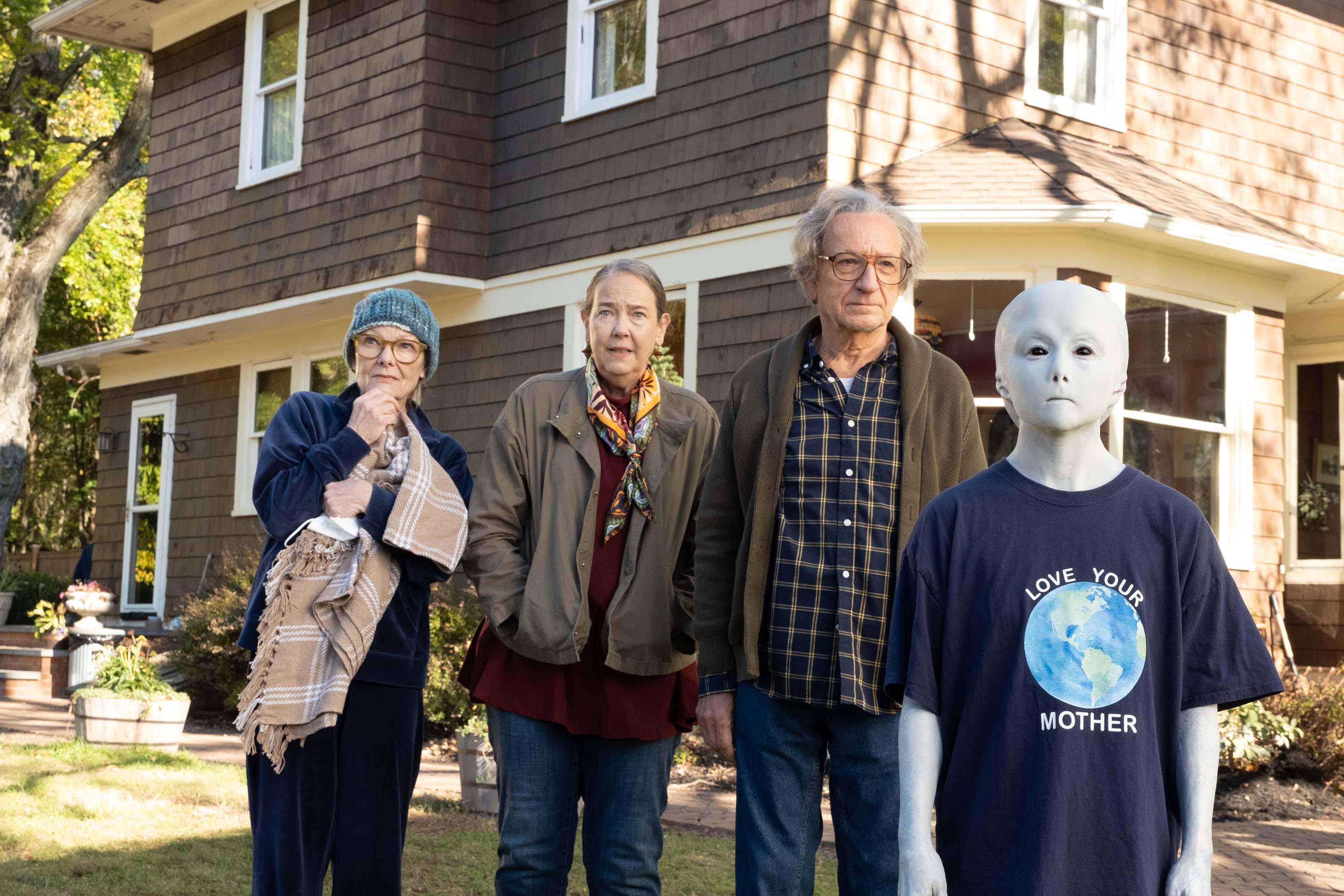 An older man and two older woman stand behind a white alien wearing a t-shirt with an Earth on it in Jules