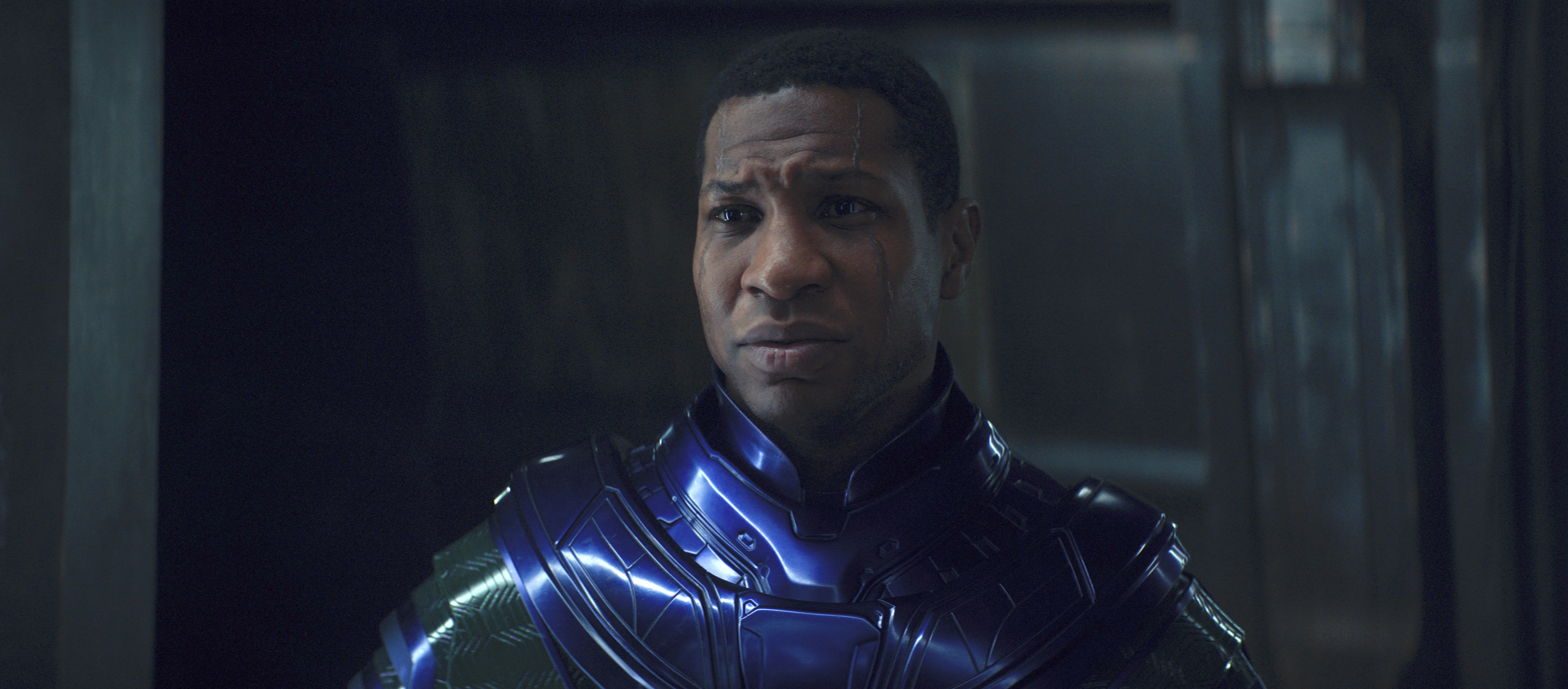 Jonathan Majors as Kang the Conqueror in Ant-Man and the Wasp: Quantumania. He wears a shiny, engraved power suit and sad. His face has two scars that run from from hairline to eyebrow and then cheek to jaw.
